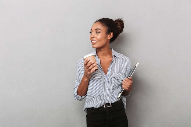 Jolie jeune femme d'affaires africaine portant une chemise debout isolée, tenant une tablette, boire du café à emporter