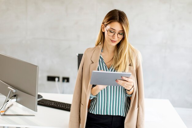 Jolie jeune femme d'affaires tenant une tablette numérique et debout dans le bureau moderne