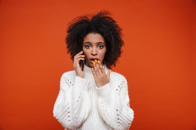 Jolie jeune femme africaine confuse portant un pull parlant au téléphone portable isolé sur un mur rouge