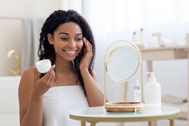 Jolie jeune femme afro-américaine appliquant une crème hydratante sur le visage