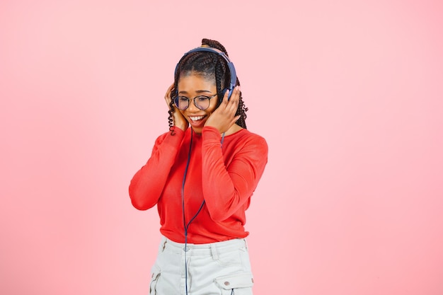 Jolie jeune femme afro-américaine avec un sourire éclatant vêtue de vêtements décontractés, des lunettes et des écouteurs dansant sur fond rose