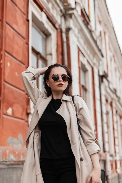 Jolie jeune femme américaine aux cheveux bruns en trench-coat élégant posant debout à l'extérieur de la ville. Le mannequin urbain belle fille élégante marche dans la rue. Vêtements d'extérieur tendance pour femmes de saison.