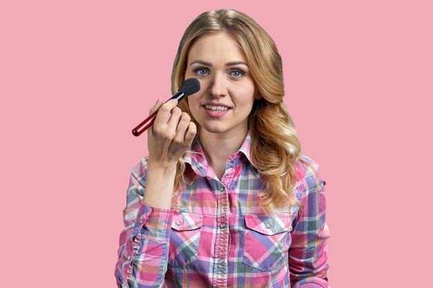 Jolie jeune femme appliquant un pinceau de maquillage sur son visage isolé sur rose