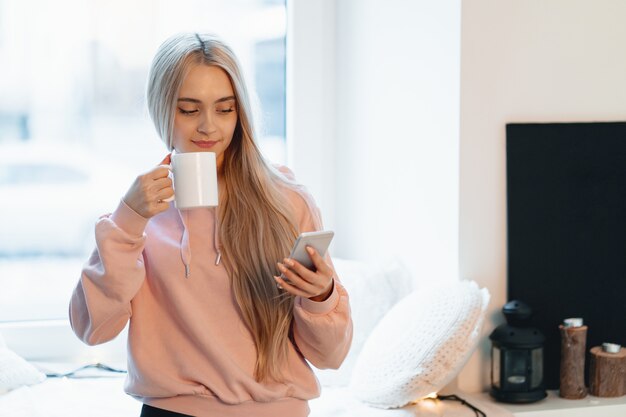 Jolie jeune femme appréciant le café tout en utilisant un smartphone par les fenêtres à l'intérieur