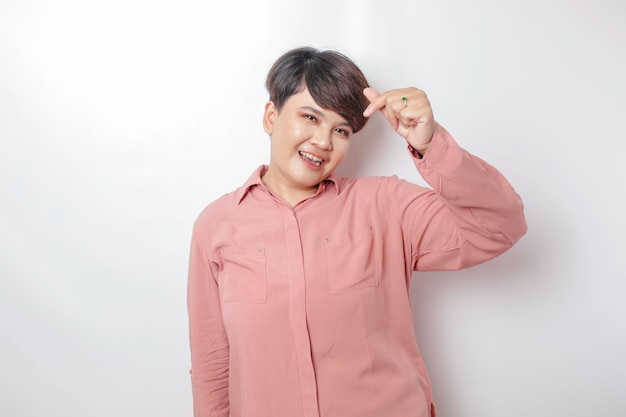 Une jolie jeune femme asiatique aux cheveux courts portant une chemise rose se sent heureuse et un geste de cœur aux formes romantiques exprime des sentiments tendres