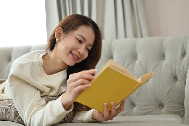 Jolie jeune femme asiatique lisant un livre ou son roman préféré sur le canapé Concept de style de vie