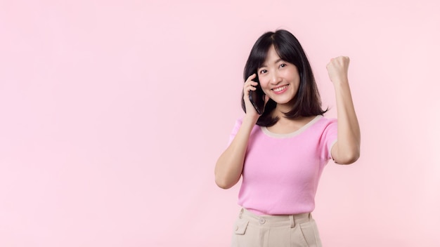 Jolie jeune femme asiatique montrant le geste de la main de la victoire du succès tout en recevant d'excellentes nouvelles du smartphone sur fond rose Bonne technologie téléphone mobile publicité concept d'application en ligne