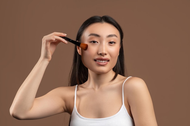 Jolie jeune femme asiatique avec un pinceau de maquillage appliquant du fard à joues sur ses joues souriant à la caméra sur