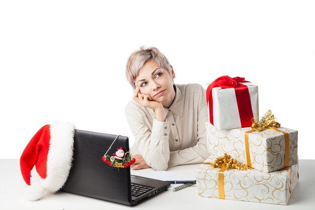 Jolie jeune femme assise au bureau avec un ordinateur portable et des boîtes à cadeaux et un chapeau d'hiver sur fond blanc