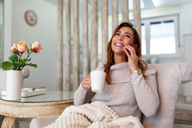 Jolie jeune femme assise sur un canapé et savourant le premier café du matin dans un appartement confortable. Jolie jeune femme discutant avec des amis sur un réseau social tout en étant assise sur un canapé à la maison et en buvant du café