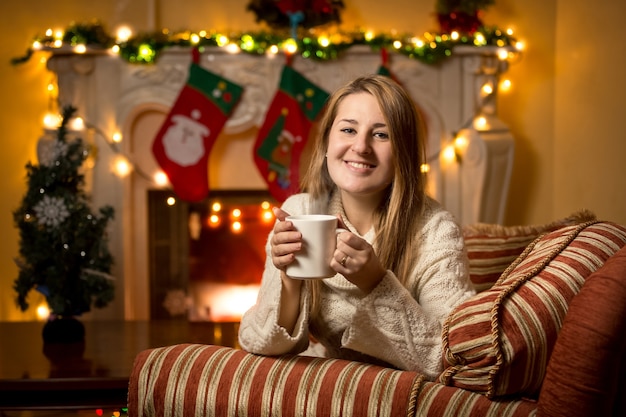 Jolie jeune femme assise à la cheminée avec une tasse de thé à Noël