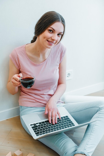 Jolie jeune femme assise près du mur sur le sol avec une tasse de café et tenant un ordinateur portable sur ses genoux