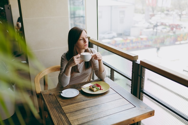 Jolie jeune femme assise seule près d'une grande fenêtre dans un café à table avec une tasse de cappuccino, un gâteau, se relaxant au restaurant pendant le temps libre. Jeune femme se reposer au café. Concept de mode de vie.