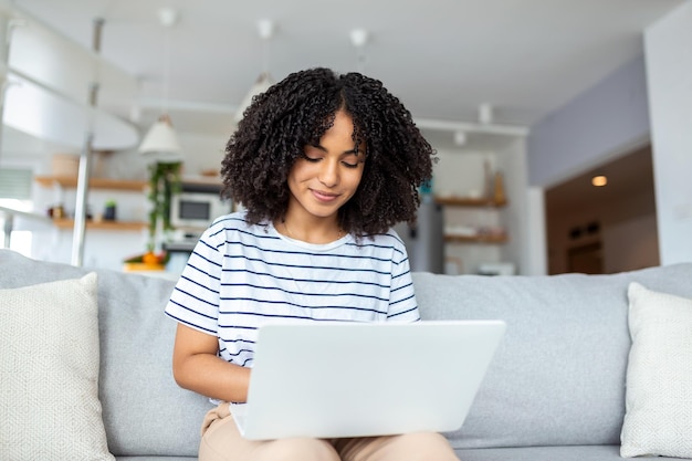 Jolie jeune femme assise seule sur son canapé à la maison et utilisant son ordinateur portable