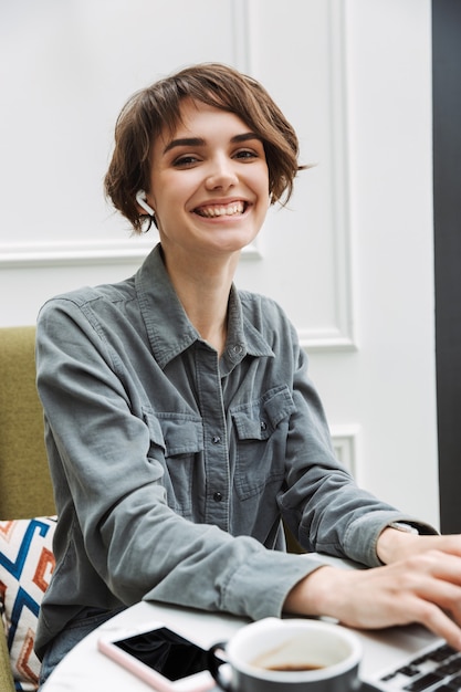 Jolie jeune femme assise à la table du café à l'intérieur, travaillant sur un ordinateur portable, analysant des documents