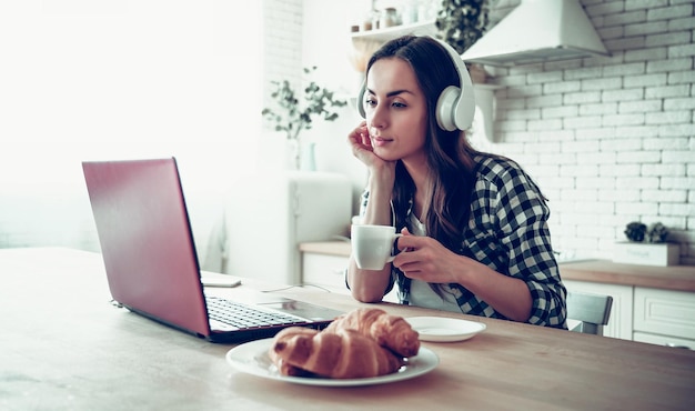 Jolie jeune femme au casque regarde un film sur un ordinateur portable dans la cuisine à la maison