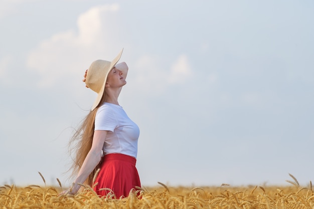 Jolie jeune femme au chapeau se tient dans un champ de blé et regarde le ciel. Vue de côté