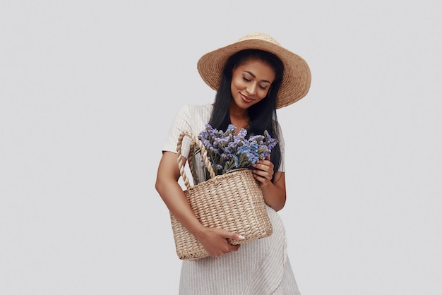 Jolie jeune femme au chapeau souriant et portant un panier avec des fleurs en se tenant debout sur fond gris