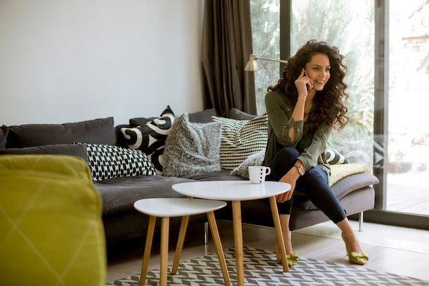 Jolie jeune femme aux cheveux bouclés à l'aide de téléphone portable par la fenêtre à la maison