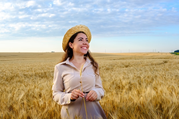 Jolie jeune femme aux cheveux longs dans un chapeau de paille tient une tige de blé dans un champ au lever du soleil et un sourire heureux. Copier l'espace