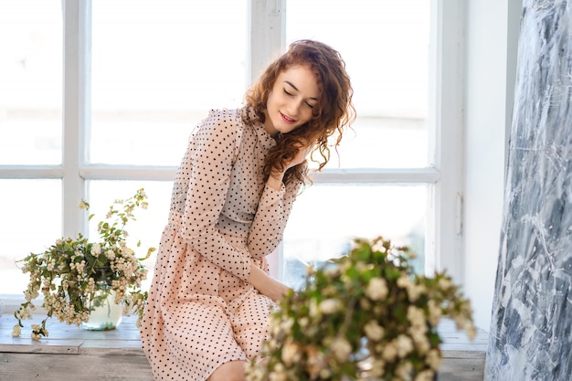 Une jolie jeune femme aux cheveux rouges dans une robe est assise sur le rebord de la fenêtre. Il y a un bouquet de fleurs à côté. Détente à la maison