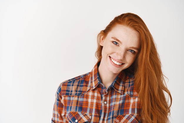 Photo jolie jeune femme aux cheveux roux et aux taches de rousseur, regardant devant avec un sourire parfait, debout en chemise contre le mur blanc