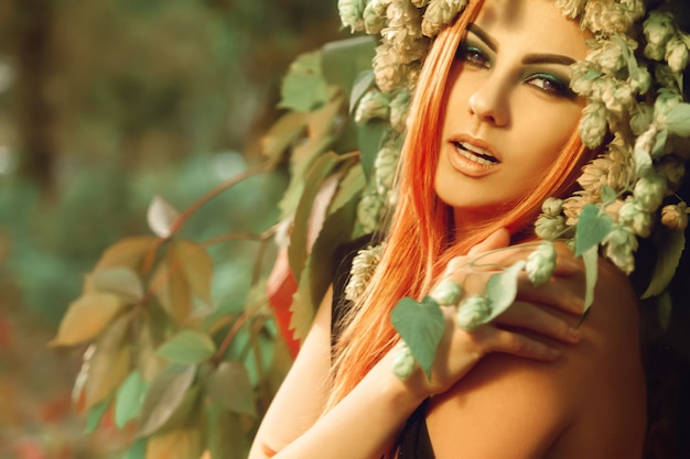 Jolie jeune femme aux cheveux roux posant avec du houblon à l'extérieur dans la forêt