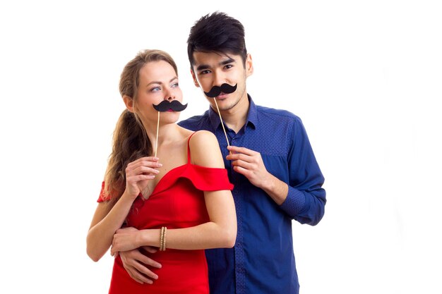 Jolie jeune femme aux longs cheveux blonds en robe rouge et jeune bel homme aux cheveux noirs en chemise bleue tenant des bâtons en carton de moustaches sur fond blanc en studio.