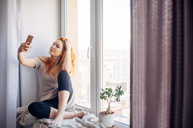 Jolie jeune femme aux longs cheveux roux prend un selfie sur son smartphone alors qu'elle est assise sur le rebord de la fenêtre. Belle fille avec un téléphone dans un environnement familial. Concept de technologie et de réseaux sociaux.