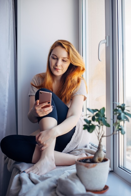 Jolie jeune femme aux longs cheveux roux utilisant un smartphone assis sur le rebord de la fenêtre à la maison, photo verticale. Concept de technologie et de réseaux sociaux.