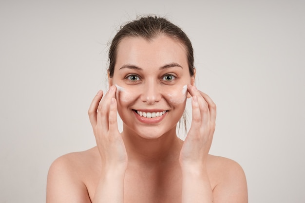 Jolie jeune femme aux seins nus souriante appliquant une crème nourrissante au visage, isoler sur un mur gris.