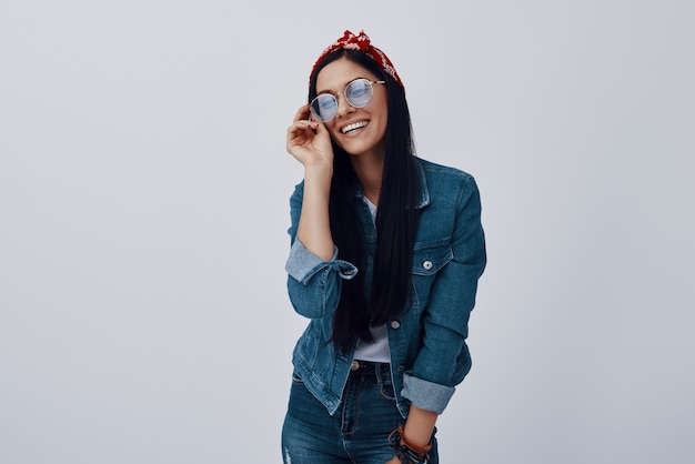 Jolie jeune femme en bandana ajustant les lunettes et souriant en se tenant debout contre le mur gris