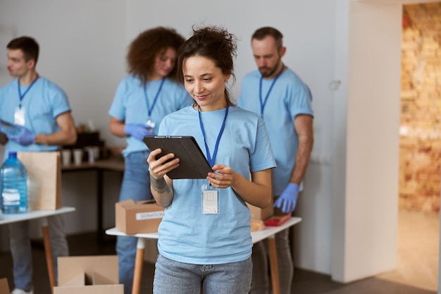 Jolie Jeune Femme Bénévole En Uniforme Bleu Utilisant Une Tablette Et Souriante En Se Tenant Debout