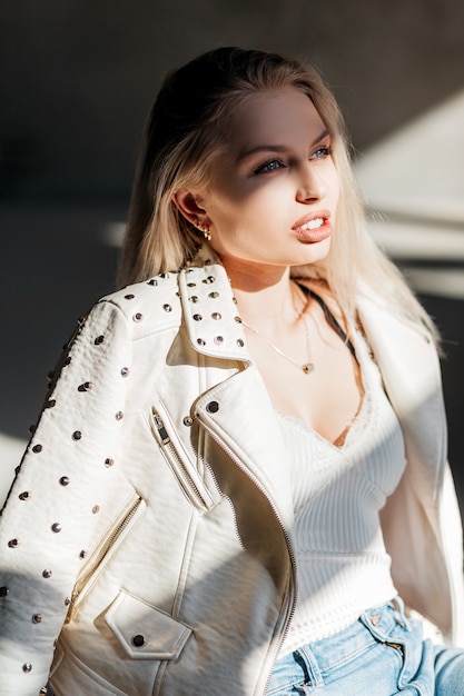 Jolie jeune femme blonde dans un T-shirt élégant blanc dans une veste d'été à la mode en blue-jeans bénéficie d'un soleil éclatant un jour de printemps