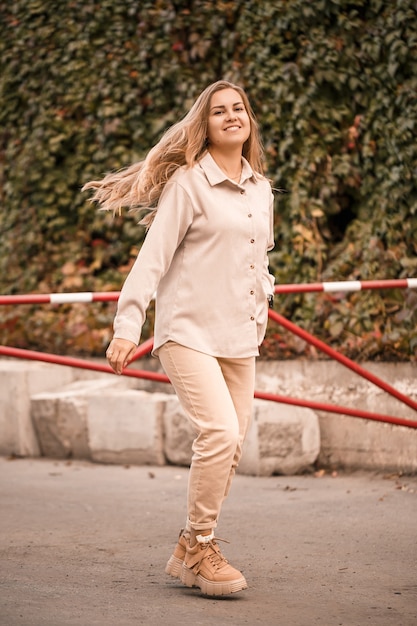 Une jolie jeune femme blonde marche dans la rue, elle porte un jean et une chemise beige. Belle fille habillée dans un style décontracté avec un sourire sur son visage pour une promenade