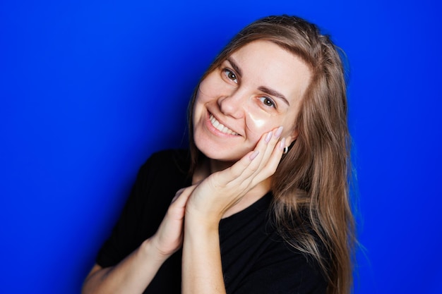 Jolie jeune femme blonde avec une peau lisse naturelle sans maquillage posant heureuse et souriante à la caméra tout en se tenant en t-shirt sur fond bleu Concept de beauté patchs oculaires pour les femmes