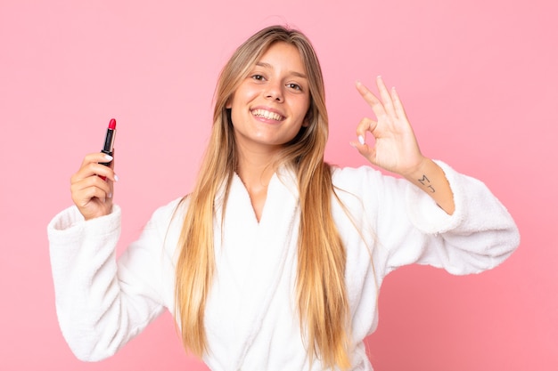 Jolie jeune femme blonde portant un peignoir et tenant un rouge à lèvres