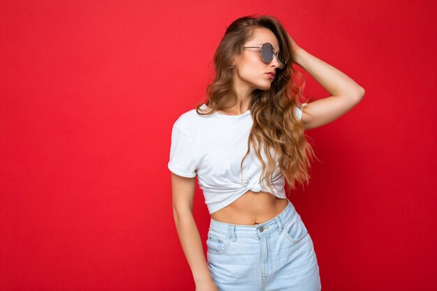 Jolie jeune femme blonde portant des vêtements élégants de tous les jours et des lunettes de soleil modernes isolées sur un mur de fond coloré regardant sur le côté