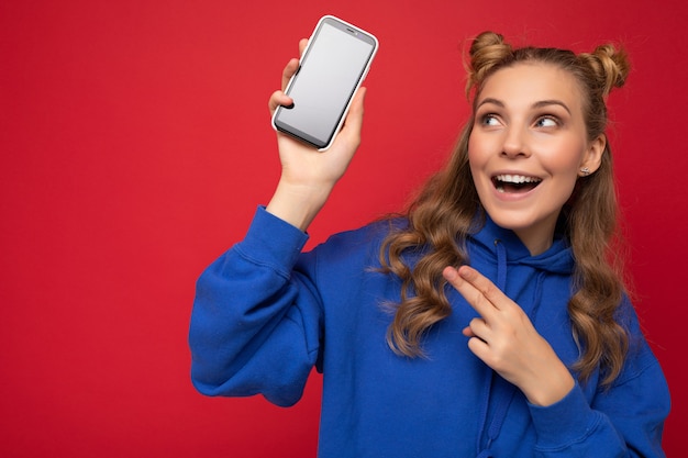 Jolie jeune femme blonde souriante portant un sweat à capuche bleu élégant isolé sur fond rouge avec