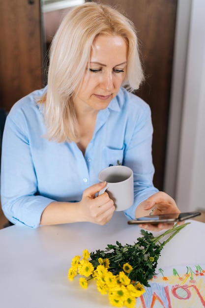 Jolie Jeune Femme Blonde Utilisant Un Téléphone Portable Et Buvant Du Café Assis à La Table Blanche. Sur Le Bureau, Des Chrysanthèmes Et Des Dessins Dessinés Par La Fille Sont Un Cadeau Pour La Fête Des Mères.