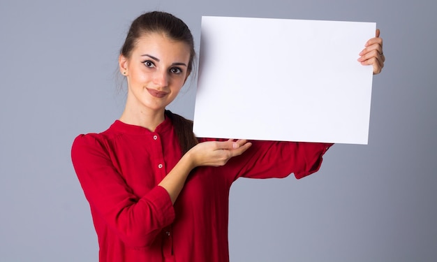 Jolie jeune femme en blouse rouge avec tresse tenant une feuille de papier blanche sur fond gris en studio