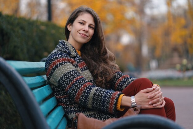 Jolie jeune femme de bonne humeur posant dans la journée d'automne, assise sur un banc, profitant du beau temps.