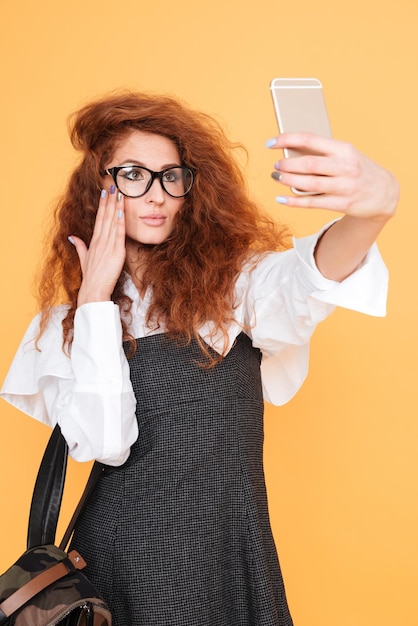 Jolie jeune femme bouclée dans des verres avec sac à dos prenant selfie à l'aide d'un smartphone