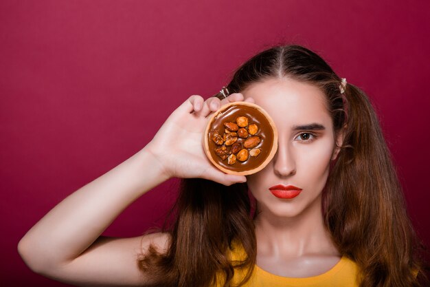 Jolie jeune femme brune aux lèvres rouges posant avec un gâteau au caramel à la main couvrant son œil. Espace libre