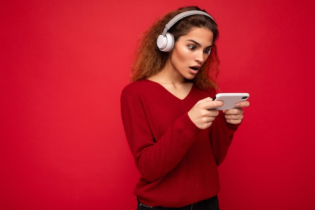 Jolie jeune femme brune bouclée choquée portant un pull rouge foncé isolé sur fond rouge portant