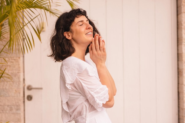 Jolie jeune femme brune caucasienne en vêtements décontractés fermant les yeux tient les paumes ensemble près du visage à l'extérieur