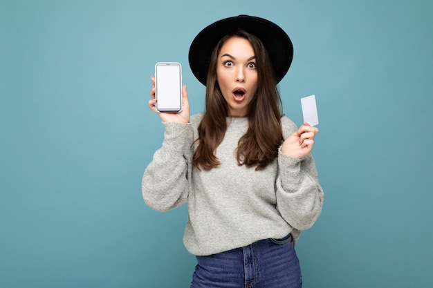 Jolie jeune femme brune choquée portant un chapeau noir et un pull gris isolé sur fond bleu tenant une carte de crédit et un téléphone portable avec un écran vide pour une maquette regardant la caméra.