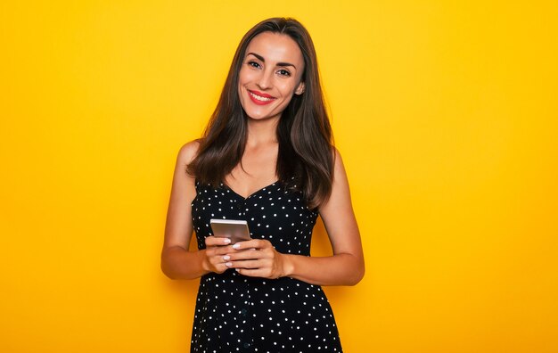 Jolie jeune femme brune confiante et moderne utilise son téléphone intelligent alors qu'elle est isolée sur un mur jaune