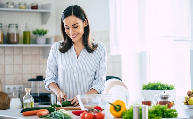 Jolie jeune femme brune heureuse de bonne humeur prépare une salade végétalienne fraîche pour une vie saine dans la cuisine de sa maison