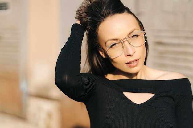 Jolie jeune femme brune portant un haut noir et des lunettes transparentes posant dans la rue. Closeup portrait de femme blanche joyeuse dans des verres